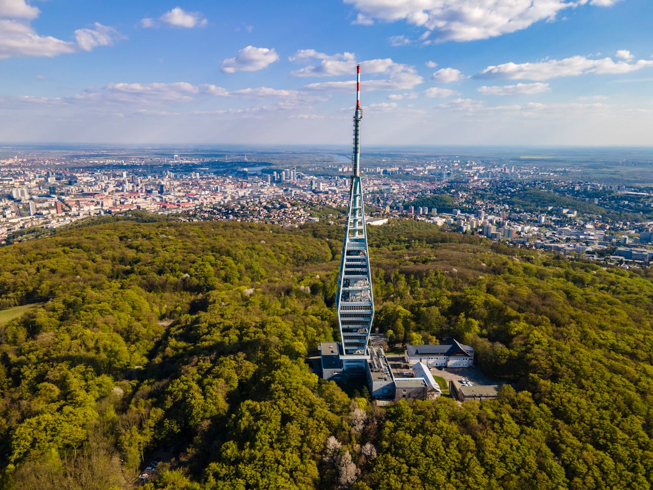 Aerial view of Kamzík TV Tower surrounded by greenery with Bratislava cityscape in the background.
