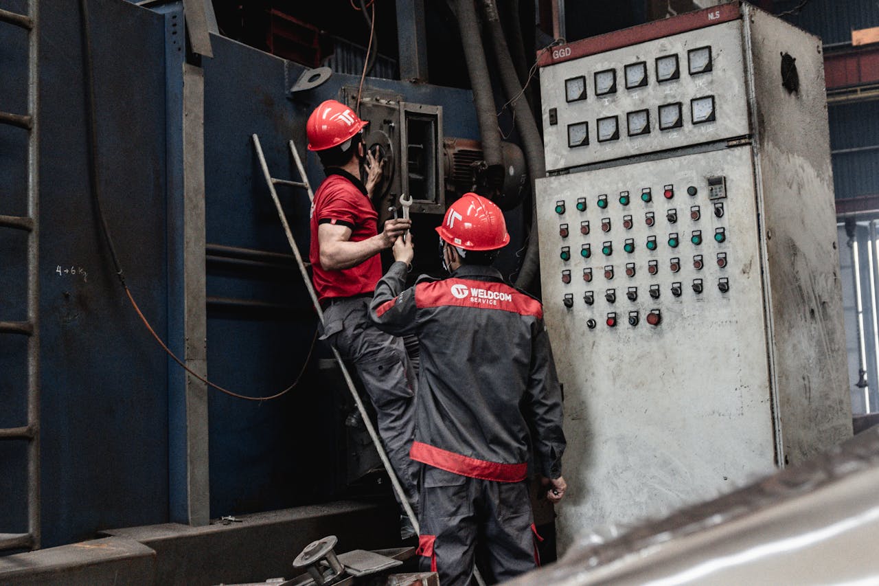 Two male workers in safety gear servicing industrial machinery inside a factory.