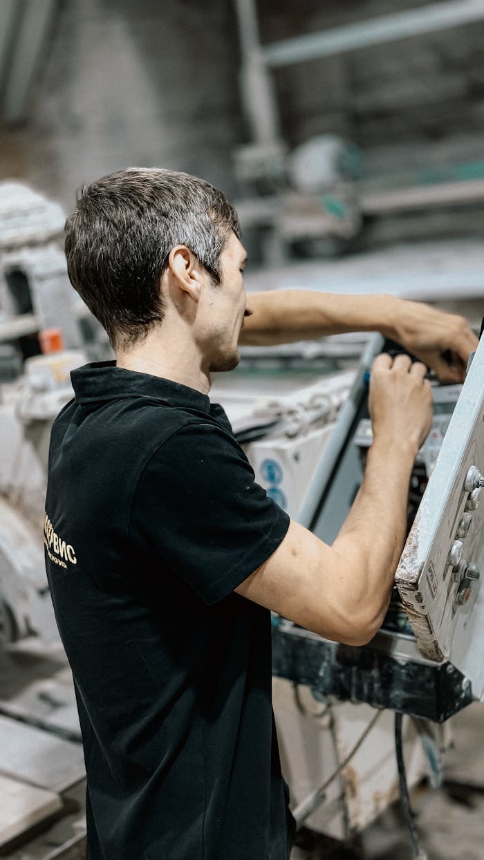 A man in a black shirt fixing machinery in an industrial workshop setting.